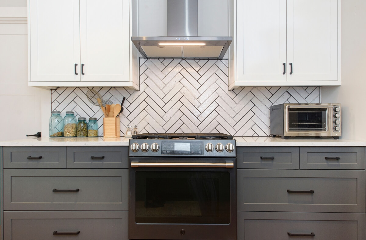 A new kitchen remodel with white painted upper cabinets (wall cabinets) and dark gray painted lower cabinets (base cabinets) with a dynamic white with dark gray grouted herringbone backsplash and modern stainless steel appliances.