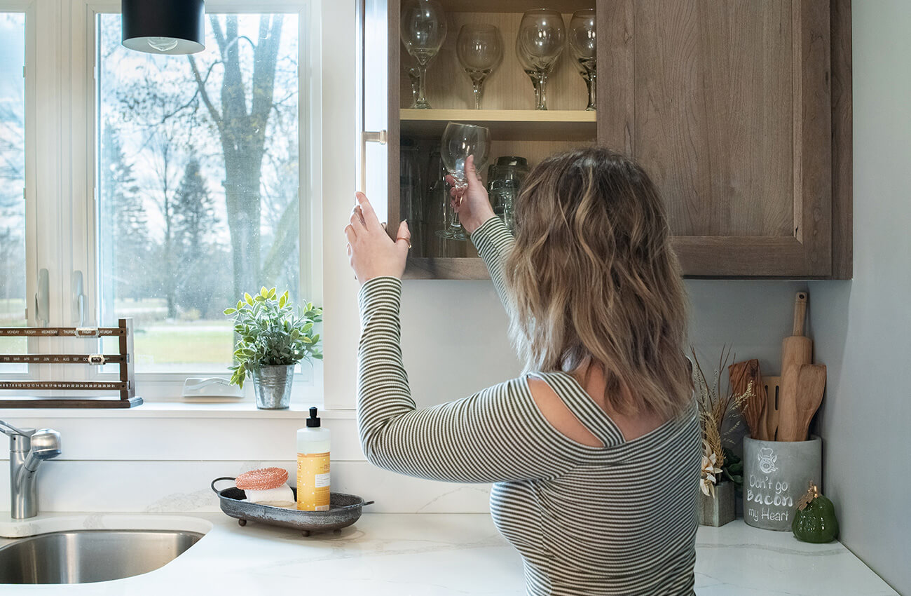 A homeowner in a newly remodeled kitchen taking a glass from a wall cabinet. The medium stained cherry cabinets are solid wood.