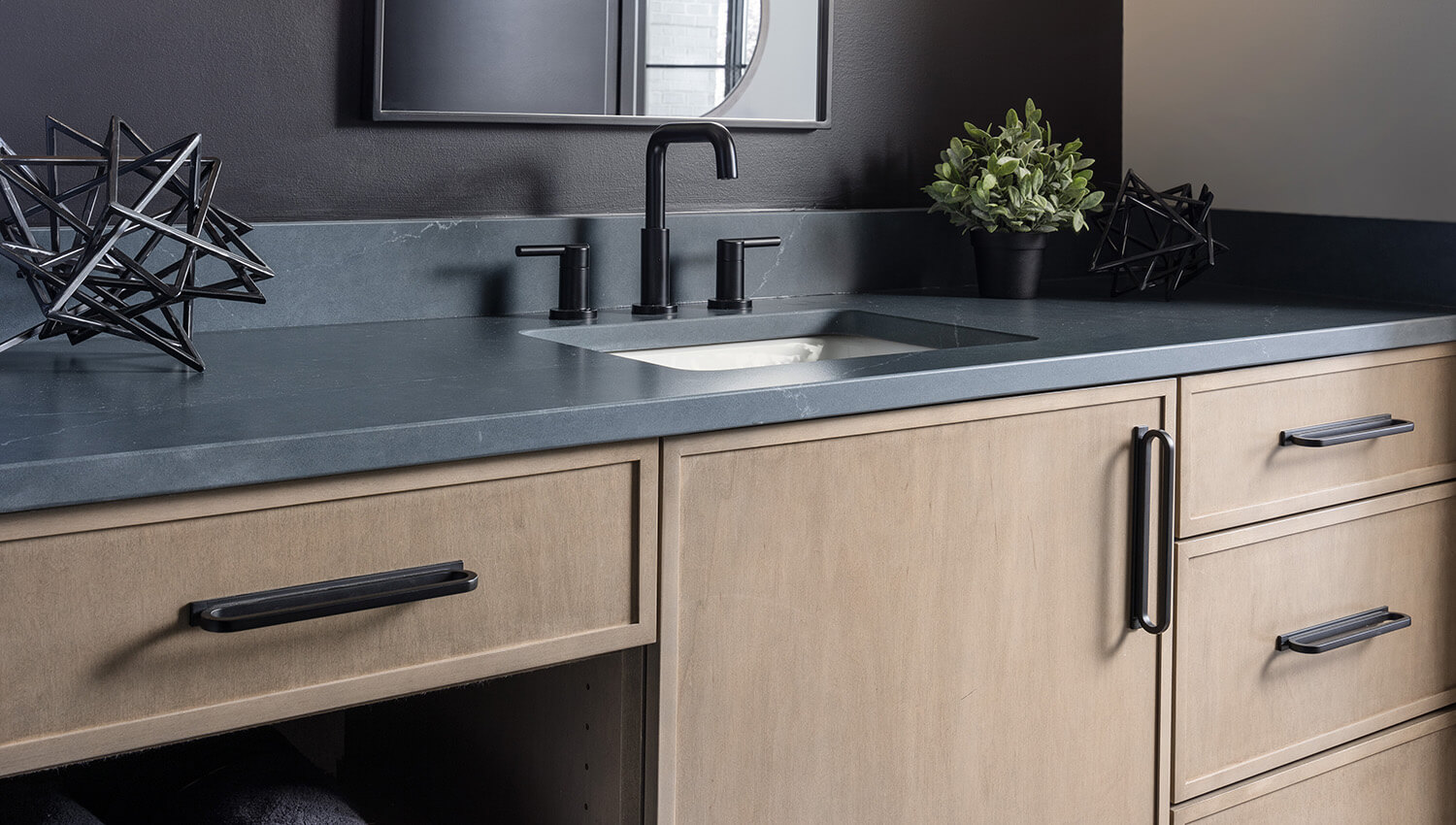A close up of a bathroom vanity in a light true-brown stain color on maple featuring a modern skinny shaker door style paired with dark gray and black finishes.