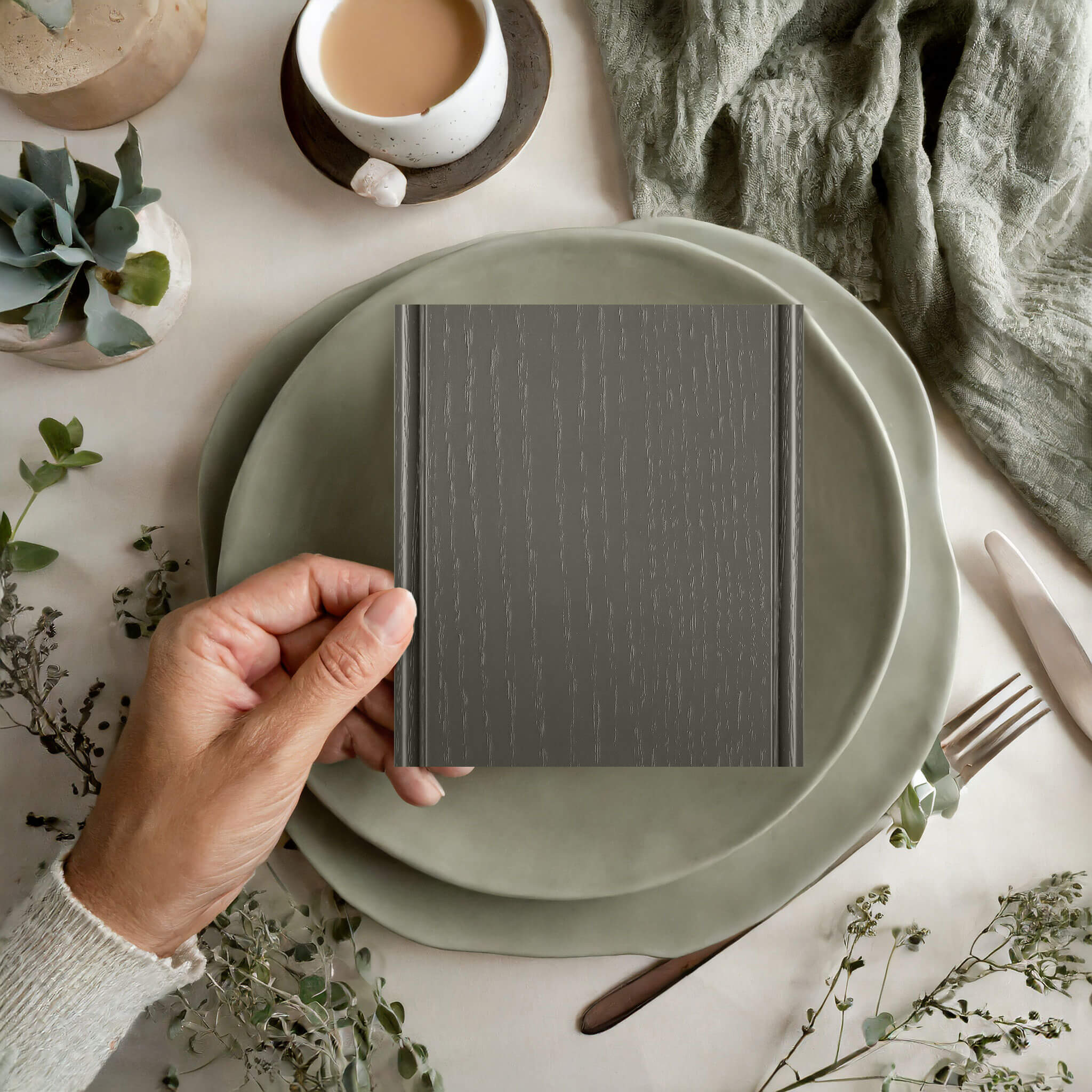 A kitchen design mood board with a hand holding a sample of painted oak in a earthy green painted finish with a defined wood grain texture.