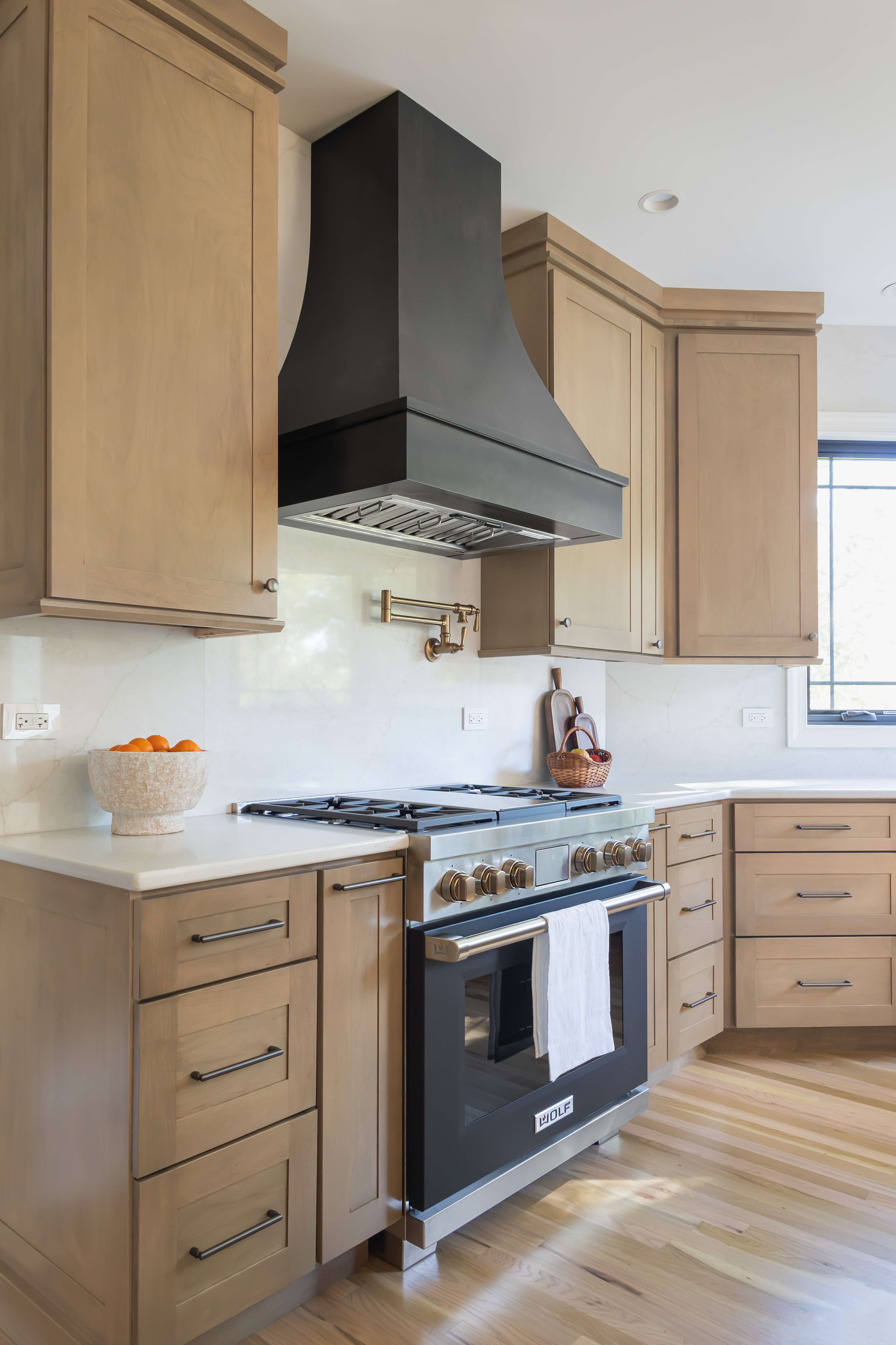 Modern shallow shaker custom kitchen cabinets in a light stained wood finish create a warm, contemporary focal point around a dramatic curved black range hood. A mix of sleek cabinet pulls and coordinating knobs adds visual interest while reinforcing the clean, tailored design.