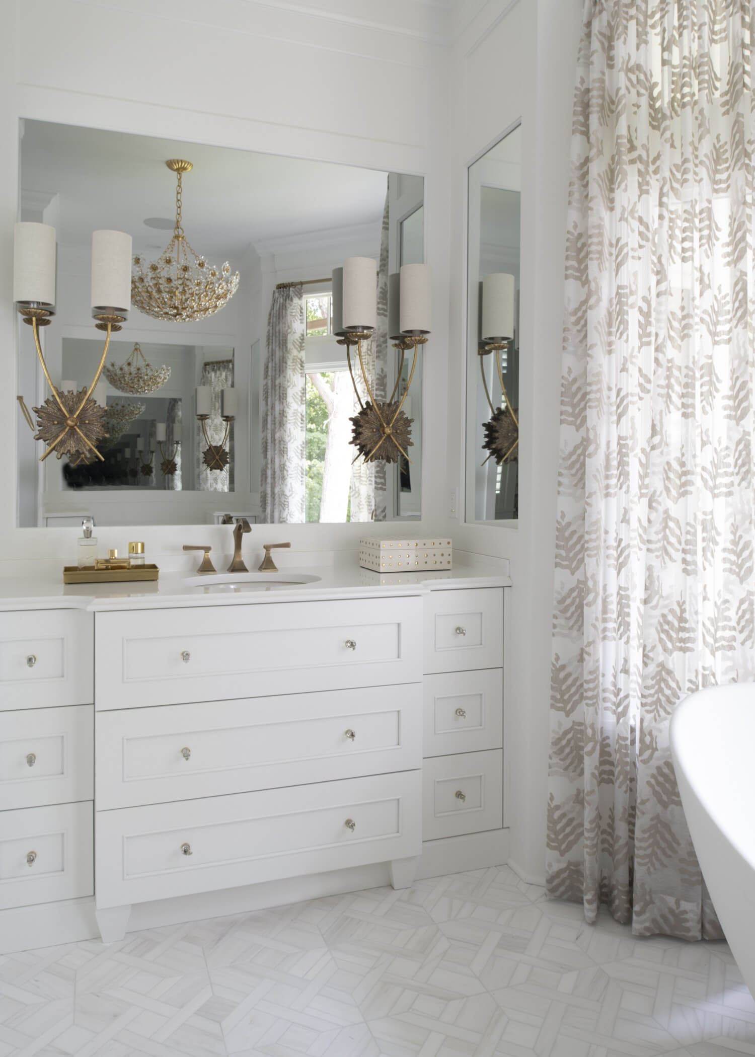 White-on-white custom bathroom vanity creates a bright, airy retreat with a furniture-style design, abundant drawer storage, and polished chrome cabinet hardware. Mirrored walls, layered lighting, and crisp white finishes enhance the timeless elegance of this luxurious bathroom.