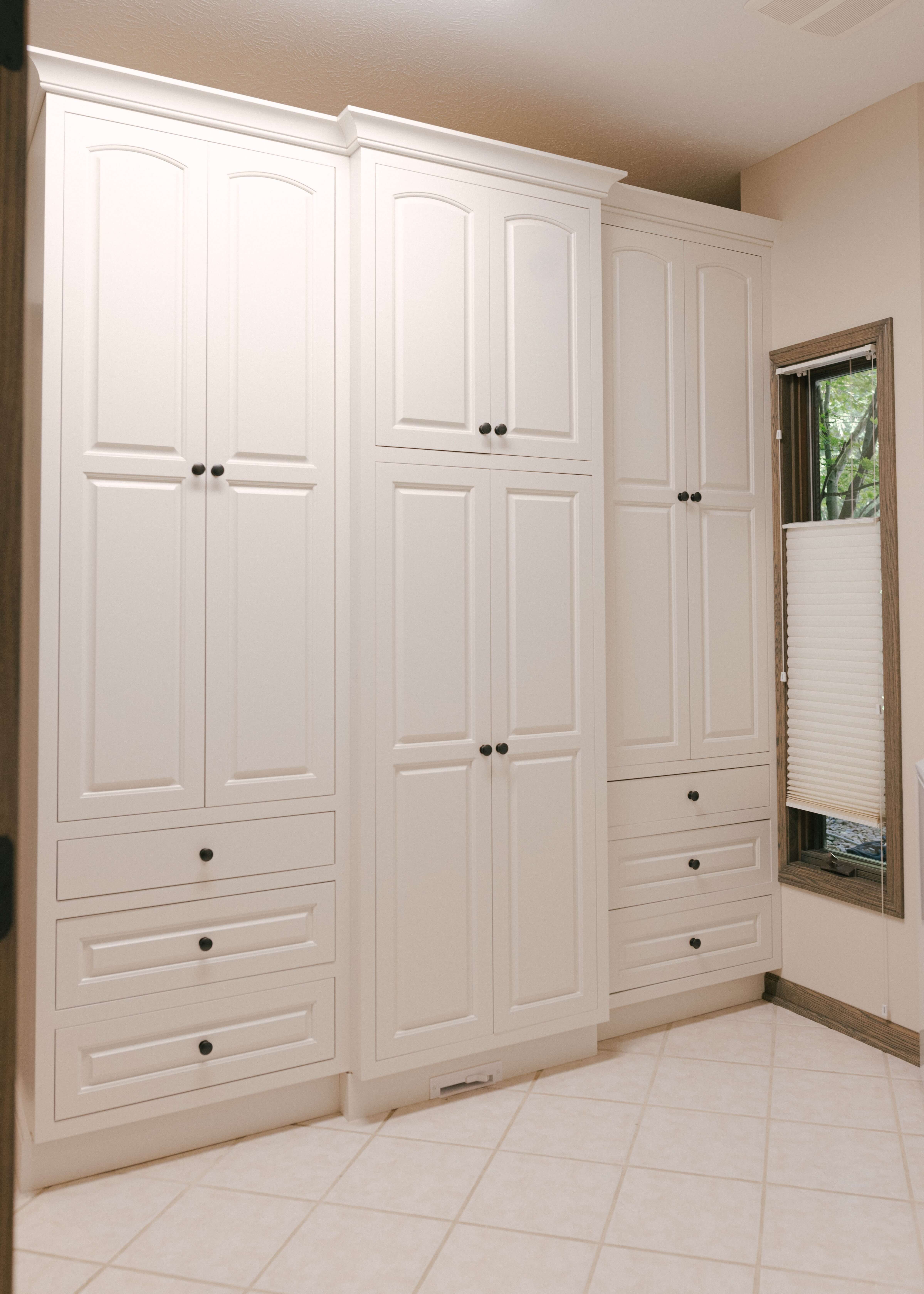Built in cabinets create a full wall of storage with tall custom cabinetry, integrated drawers, and overhead compartments for maximum organization. White painted cabinetry and streamlined hardware deliver a timeless, tailored look.